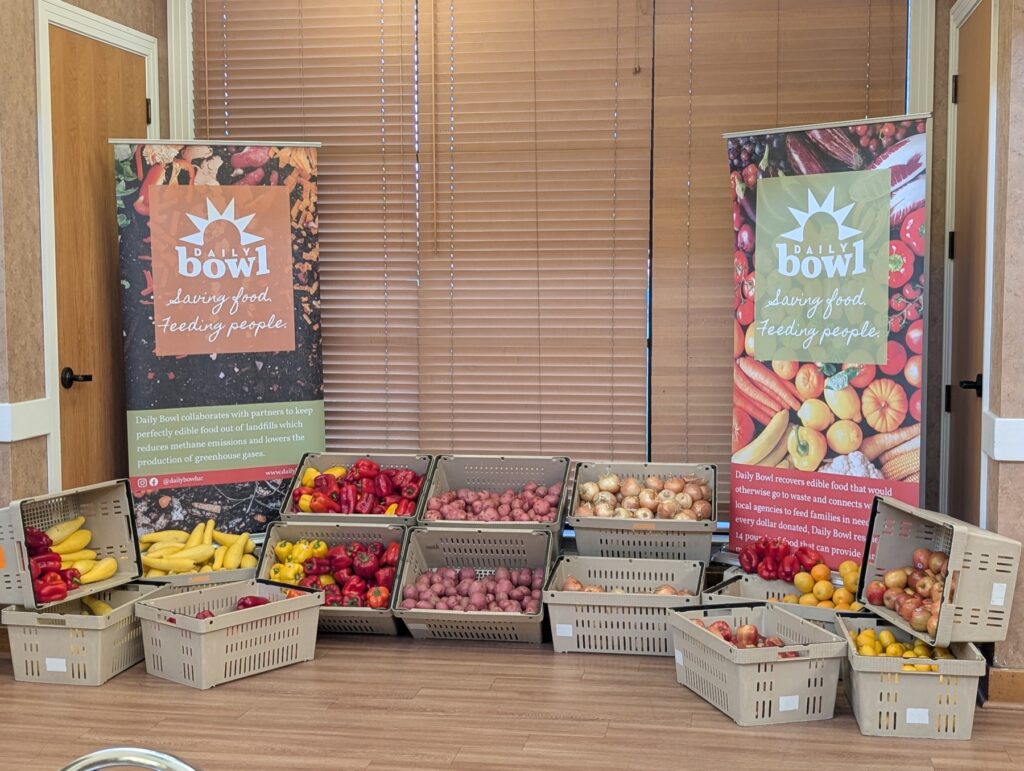Crates of rescued peppers, onions, bananas and apples between two Daily Bowl banners