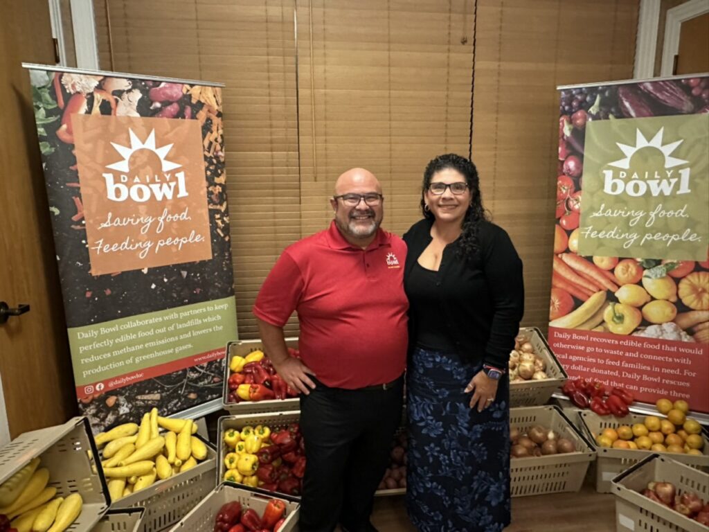 Two of the team in front of a Daily Bowl banner and crates of produce
